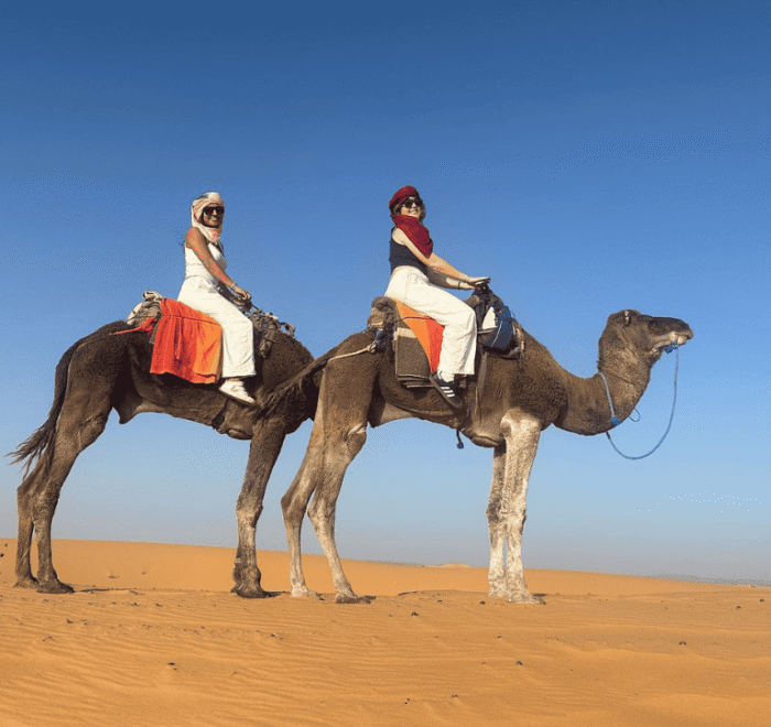 Two people in headscarves and sunglasses ride camels through a sandy desert under a clear blue sky. Perfect for those seeking Morocco holiday packages 2026, the riders pose for a photo atop camels with red-blanketed saddles.