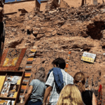 Four people browse colourful textiles, photos, and crafts displayed on a rustic stone wall under a bright sky, with ancient ruins visible above in the background.