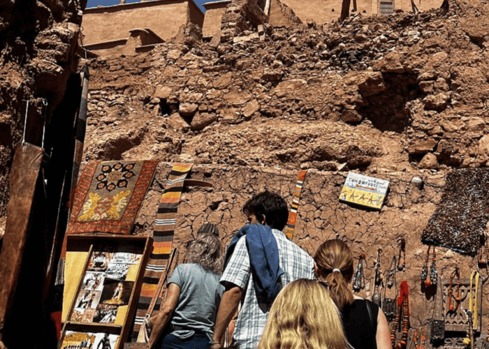 Four people browse colourful textiles, photos, and crafts displayed on a rustic stone wall under a bright sky, with ancient ruins visible above in the background.