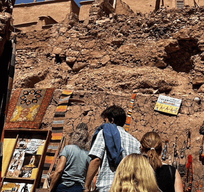 Four people browse colourful textiles, photos, and crafts displayed on a rustic stone wall under a bright sky, with ancient ruins visible above in the background.