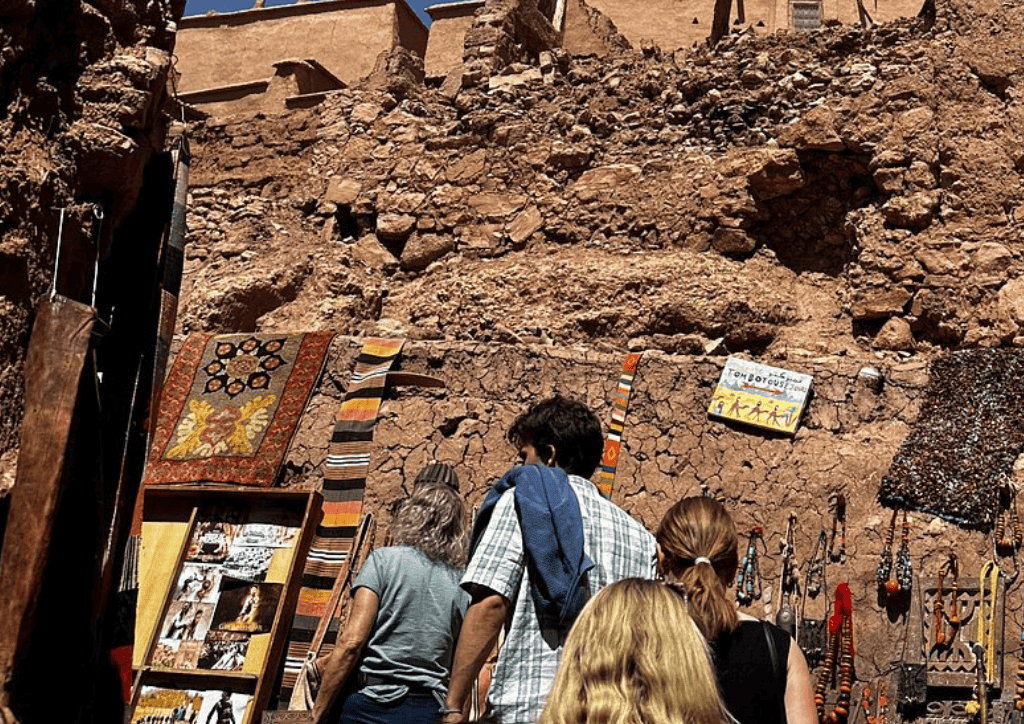 Four people browse colourful textiles, photos, and crafts displayed on a rustic stone wall under a bright sky, with ancient ruins visible above in the background.
