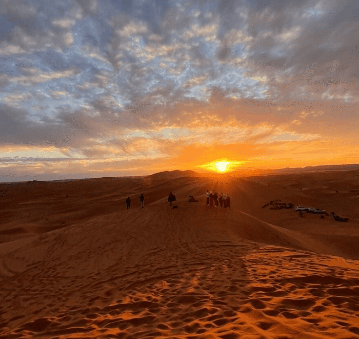 People stand atop sand dunes watching a vibrant sunset, with golden rays lighting up the sky and clouds. The vast desert landscape, perfect for Morocco holiday packages 2026, stretches out under dramatic, colourful clouds.