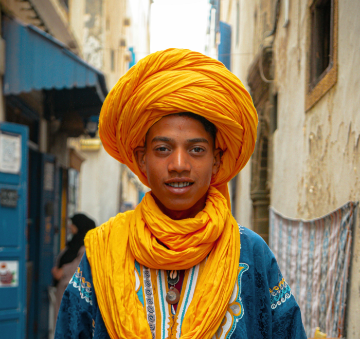 A young person in a vibrant yellow turban and scarf, wearing a blue embroidered garment, stands smiling in a sunlit alley lined with old buildings—capturing the spirit of Morocco holiday packages 2026.