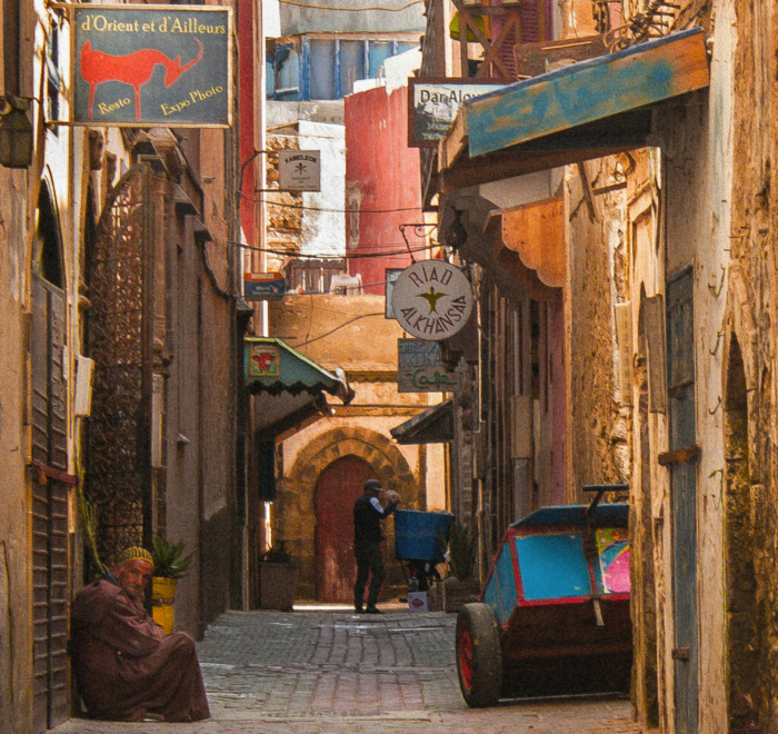 A narrow cobbled street in a historic town features colourful shop signs, carts, and a person in traditional clothing sitting by the wall. Two people stand near an arched doorway in the background. Warm sunlight bathes this Morocco holiday packages 2026 scene.