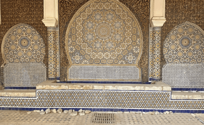 Intricate Moroccan mosaic tilework with blue, white, and gold geometric patterns decorates a large arched wall fountain. The ornate designs and Arabic calligraphy evoke the timeless beauty featured in Morocco holiday packages 2026.