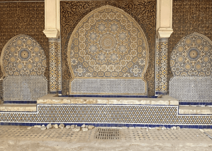 Intricate Moroccan mosaic tilework with blue, white, and gold geometric patterns decorates a large arched wall fountain. The ornate designs and Arabic calligraphy evoke the timeless beauty featured in Morocco holiday packages 2026.