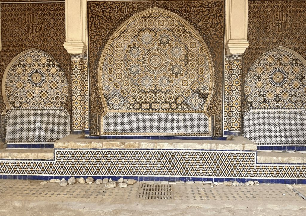 Intricate Moroccan mosaic tilework with blue, white, and gold geometric patterns decorates a large arched wall fountain. The ornate designs and Arabic calligraphy evoke the timeless beauty featured in Morocco holiday packages 2026.